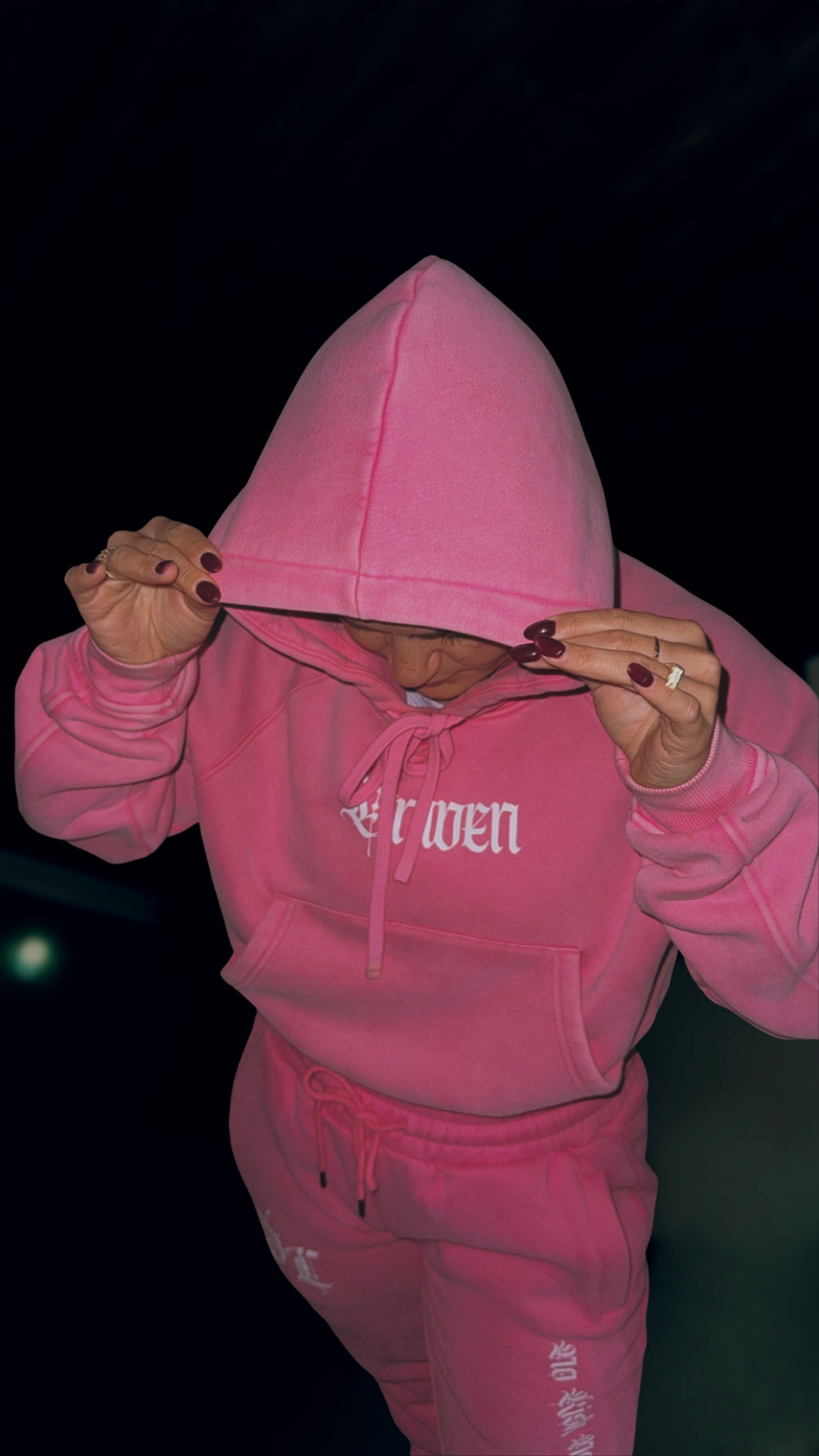 Person wearing a pink hoodie and pants with text, holding the hood against a dark background