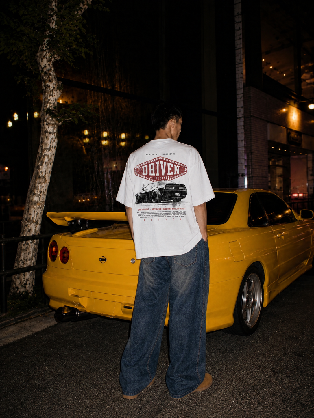 Guy posing in front of a yellow car wearing a white Driven Lifestyle branded tee