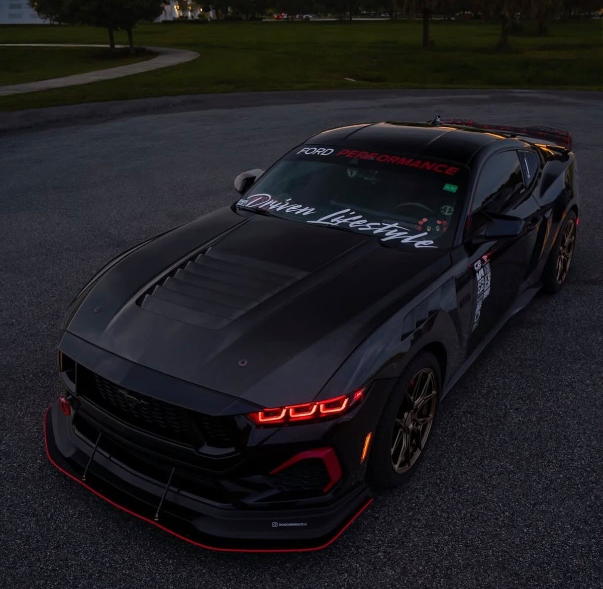 A black Ford Mustang performance car with custom decals and a front splitter, photographed on an outdoor paved surface at dusk.