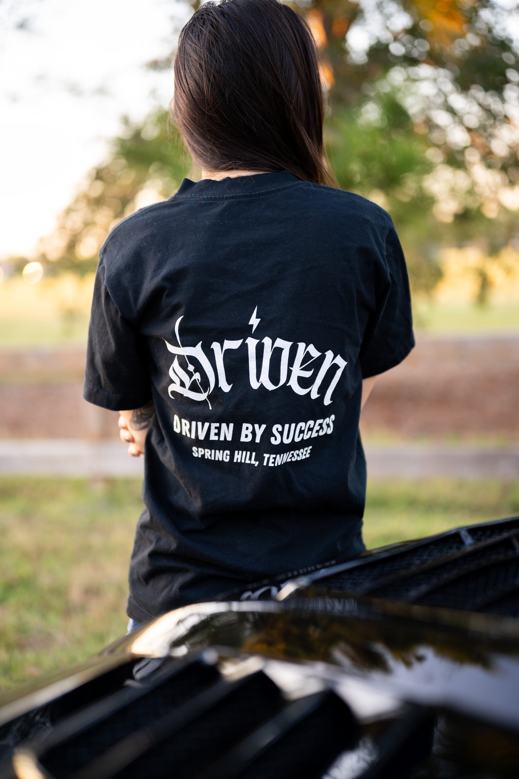 Person wearing a black t shirt with 'Driven by success spring hill, tennessee' branding outdoors, burred background