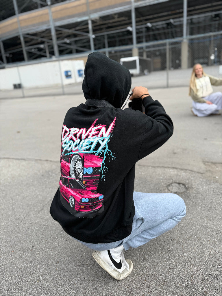 Person wearing a black hoodie with a colorful graphic and text, taking a photo in an urban setting.