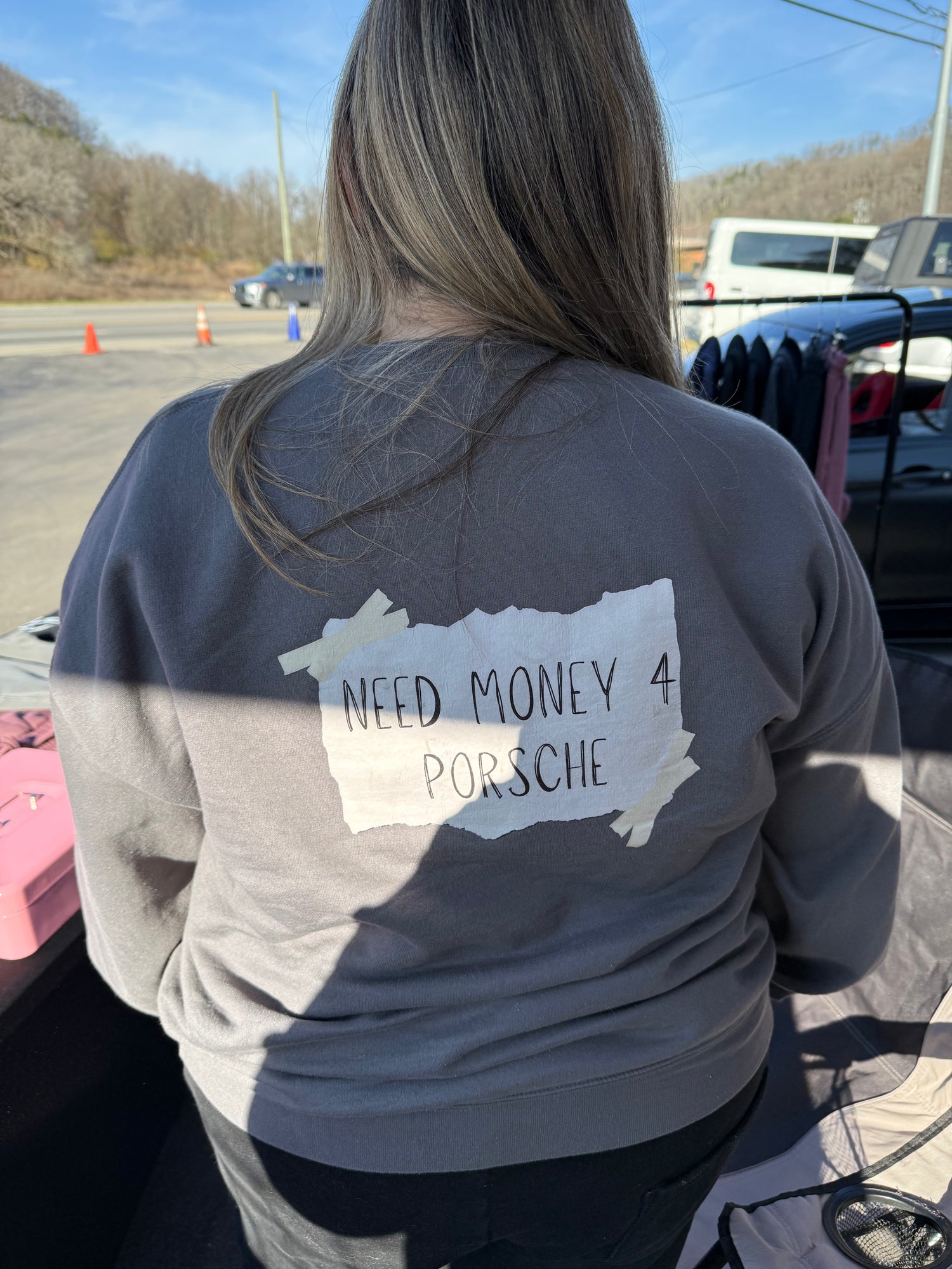 Person wearing a sweatshirt with 'Need Money 4 Porsche' text in a parking lot.