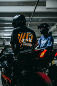 A person wearing a black motorcycle helmet and a black t-shirt with a large orange and white graphic on the back sits on a black sport motorcycle in a dimly lit parking structure. Another person in a blue shirt and black helmet is visible in the background.