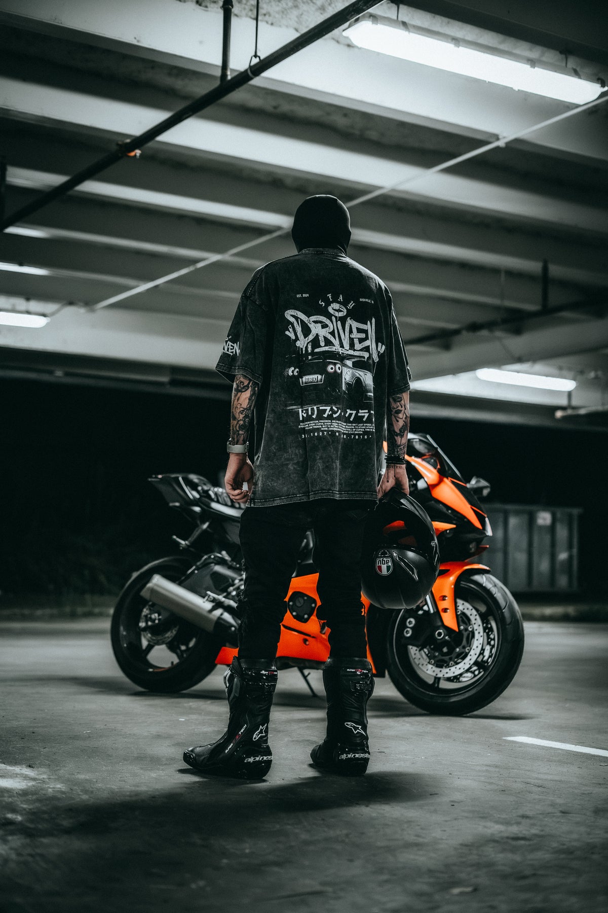 A person stands with their back to the camera in a dimly lit parking garage, wearing a black graphic t-shirt with 'DRIVEN' printed in white on the back, black pants, and black boots. An orange and black sport motorcycle is parked behind them.